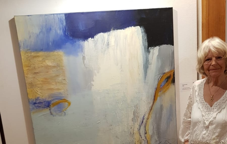 Marianne Igel standing in the exhibition to the right of a large square painting in blue, white and ochre with a yellow arc at its right edge; the exhibition label is on the wall beside it