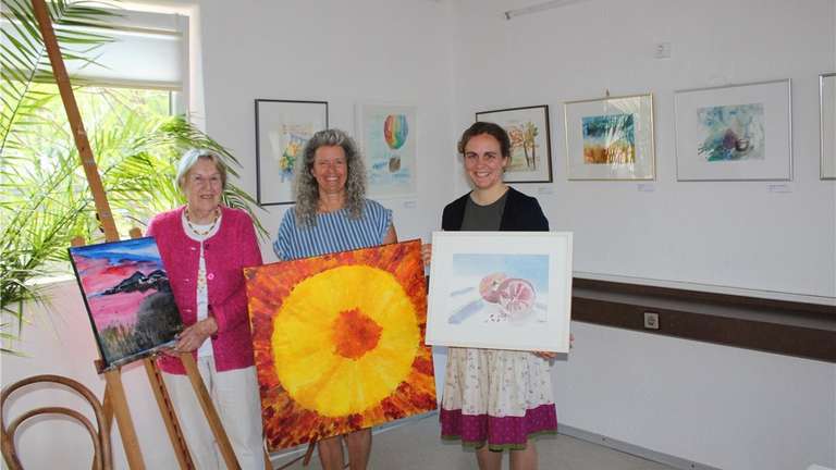 Three painters stand side by side in a bright exhibition room holding their work: folded cards and a small picture at the left, a large yellow sun form on red in the middle, and a framed watercolour in blue and pink at the right; more framed sheets hang on the walls behind them