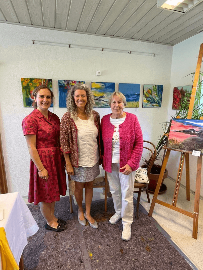 Three women stand side by side in Marianne's studio; a row of small colourful paintings hangs behind them and another stands on an easel to the right