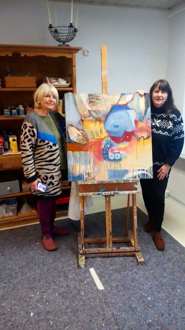 Two women stand either side of an easel in the workshop, holding a large picture in ochre, blue and red between them; behind them a wooden dresser of paint pots and jars, and the floor is flecked with paint