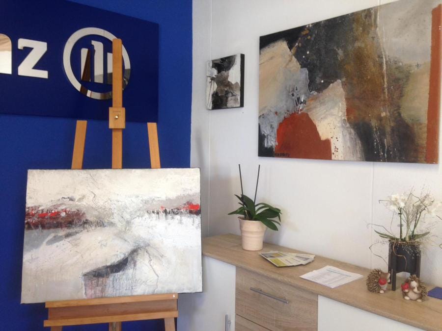 Her pictures in the insurance agency: at the left, against a blue wall carrying the Allianz mark, a painting in off-white with a red band stands on an easel; at the right, on the white wall, a small dark format and a large landscape-format picture in black, sand and rust red, with a sideboard of orchids and leaflets below
