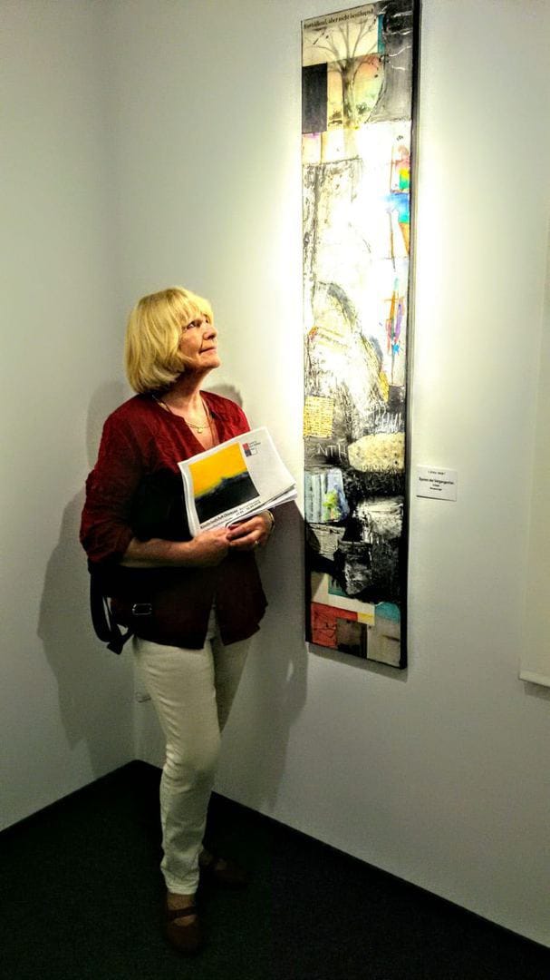 Marianne Igel stands in a corner of the exhibition room with a catalogue under her arm, looking up at a tall narrow mixed-media work hanging beside her on the white wall; the small label with title and technique is to its right