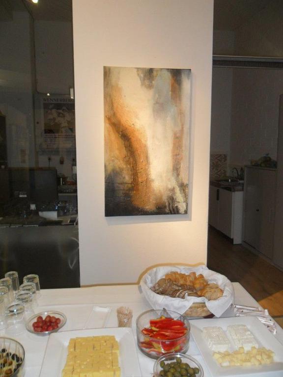 An upright picture in ochre, rust and white hangs on a white pillar; in the foreground the private view's laid table, with a bread basket, cubes of cheese, tomatoes, peppers and olives