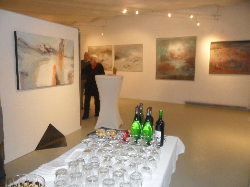A view into the gallery: several landscape-format pictures in pale earth tones on the white walls, a standing table in the middle, a table of wine bottles and many glasses in the foreground and two guests at the back; a dark pyramid stands on the floor at the left