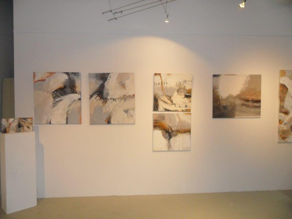 A long wall of six abstract pictures in white, sand and rust red under spotlights, with a white plinth at the left carrying a small work