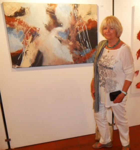 Marianne Igel stands in the exhibition beside a large landscape-format picture in rust red, white and blue; she is dressed in white with a green necklace, and another work is cut off at the right edge
