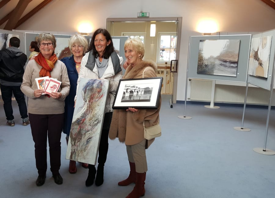 Four women stand between display panels in a bright exhibition room under a roof truss, holding works: folded cards, an upright picture in reds and greys, and a framed black-and-white photograph of a bridge; more pictures hang on the panels behind them