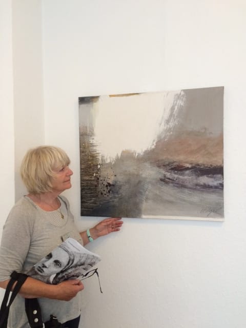 Marianne Igel stands at a white wall beside a landscape-format picture in grey, ochre and white, resting a hand on its lower edge; under her arm she holds a folder with a face printed on it