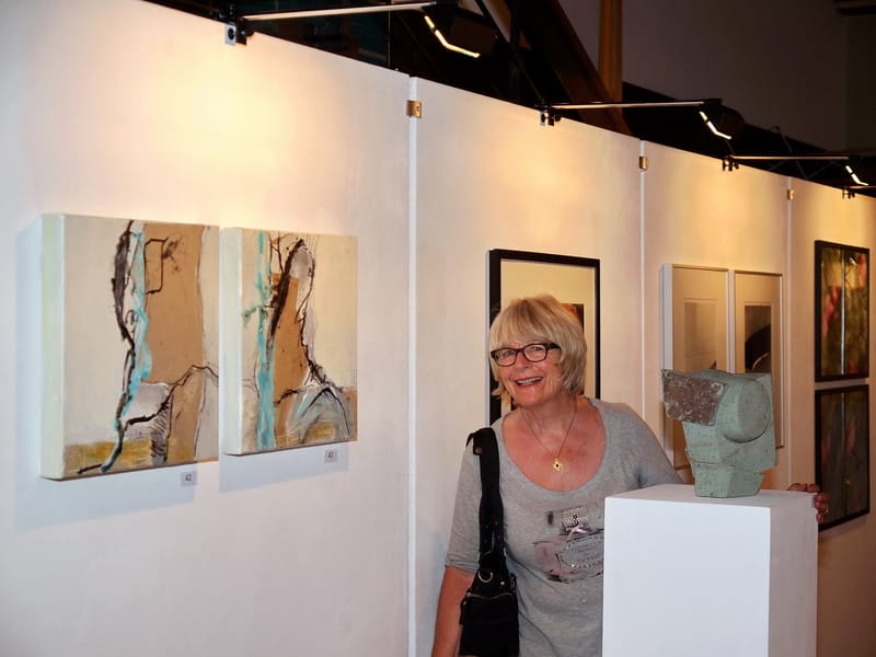 Marianne Igel stands laughing in an exhibition beside a white plinth carrying a grey-green stone sculpture; two small upright works in sand and turquoise with number tags hang on the panel at the left, and further framed works at the right