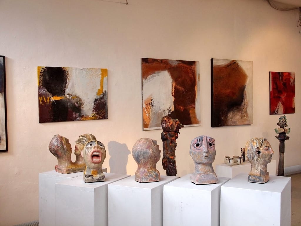 A second view of the room: a row of abstract paintings in brown, rust and red along the wall, with several ceramic heads on low white plinths in front of them