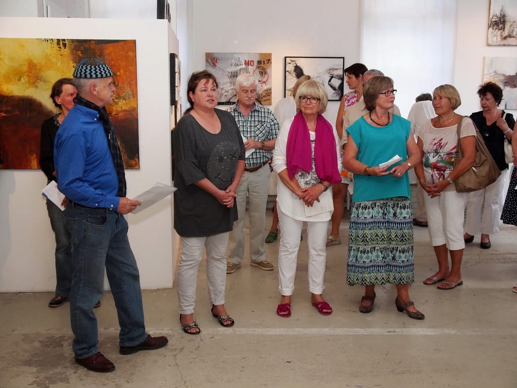 Guests stand listening in the gallery with programmes in their hands; a large orange and gold painting and other works hang behind them