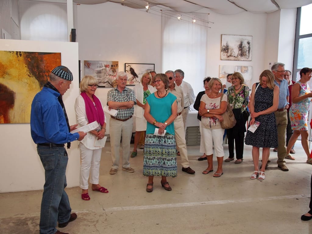 A larger group of guests stands closely together in the room at the opening, many holding a programme