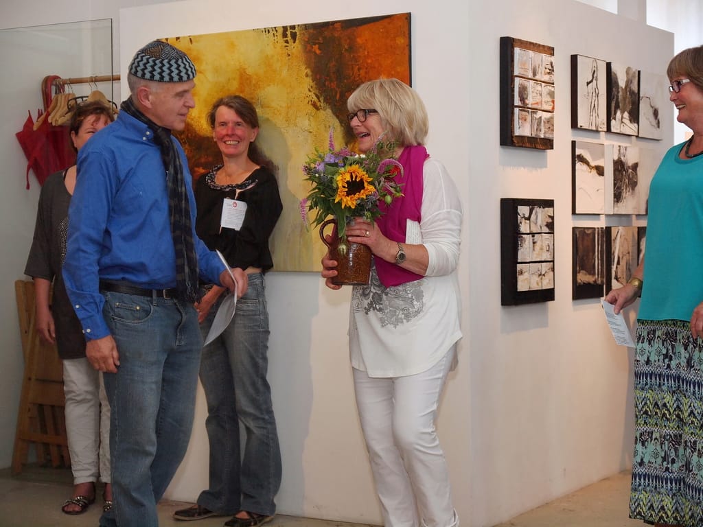 Marianne Igel laughs, holding a bunch of sunflowers in an earthenware jug; guests stand around her and a large orange and gold painting hangs behind her