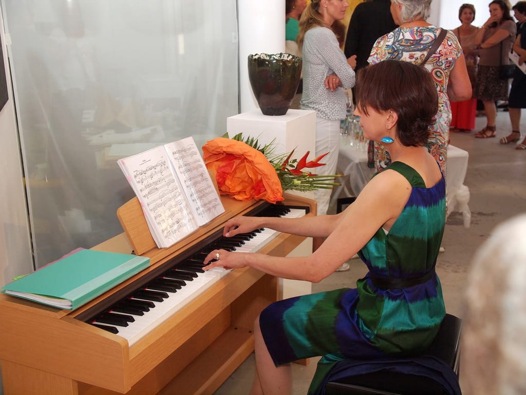 A pianist in a green and blue dress plays a wooden piano; sheet music stands on the rest with an orange bouquet beside it, and guests talk in the background
