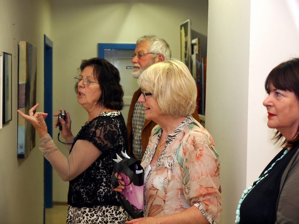 A woman points at a row of small paintings on the wall and talks about them; Marianne Igel and other guests stand beside her