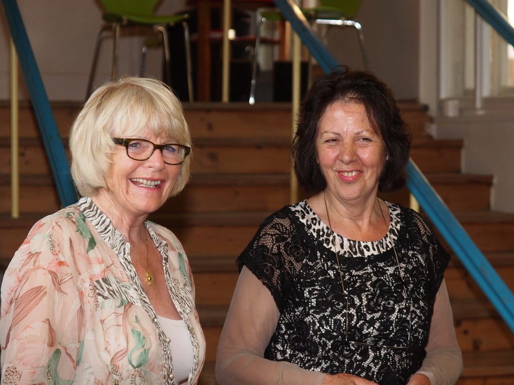 Marianne Igel and Thea Barth stand side by side, both laughing, in front of a wooden staircase with blue and yellow handrails