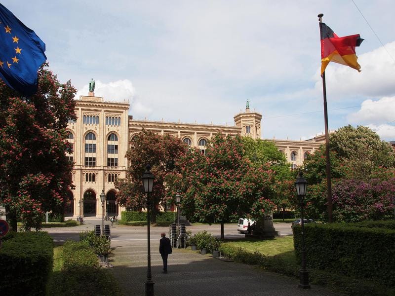 The Regierung von Oberbayern building on Maximilianstraße in Munich seen from the street: a long façade of round-arched windows and turrets, chestnut trees in flower in front of it, a European flag at the left and a German flag at the right