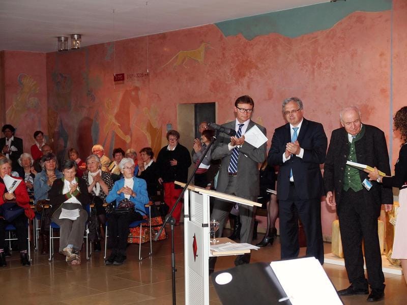 The opening in a hall painted with a pink classical mural: the audience sits on blue chairs and applauds, and three men stand at the right beside the lectern, one of them holding cards