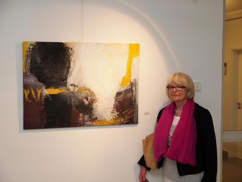 Marianne Igel stands beside her large landscape-format picture in yellow, white and dark brown on a white wall; she is wearing a pink scarf, and the small exhibition label is to the right of it